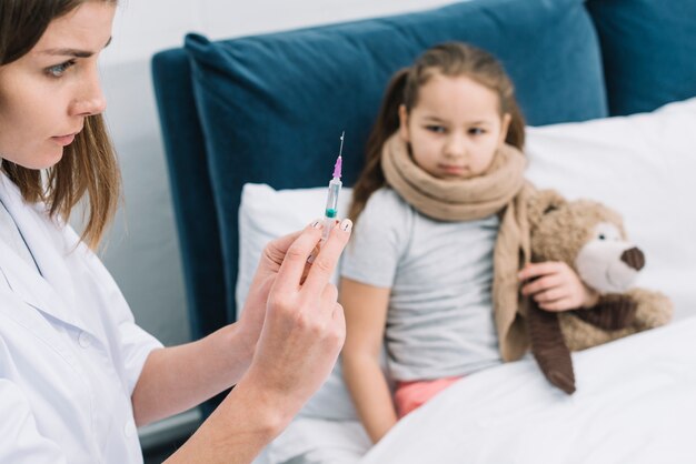 Primer plano de una doctora manos llenando la jeringa con medicina frente a una niña enferma sentada en la cama