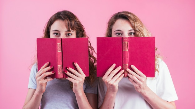 Primer plano de dos amigas sosteniendo el libro debajo de los ojos contra el fondo rosa