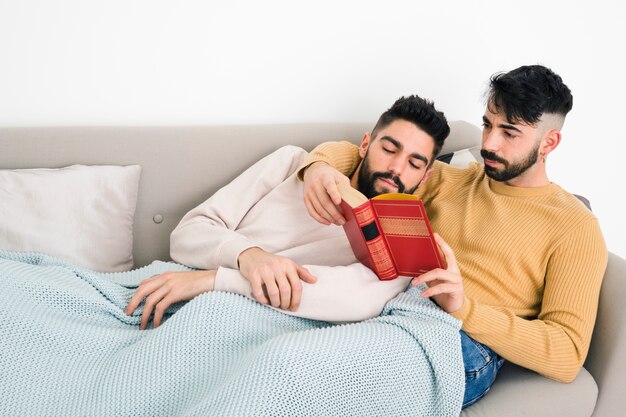 Primer plano de dos parejas homosexuales leyendo el libro acostado debajo de una manta en el sofá