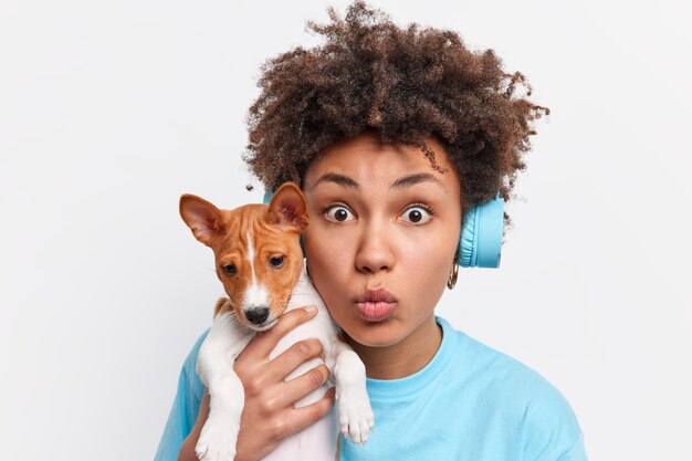 Primer plano de la dueña de una perra afroamericana lleva un cachorro de pedigrí mantiene los labios doblados y se ve sorprendido tiene el pelo rizado escucha música a través de auriculares mientras camina con su animal favorito