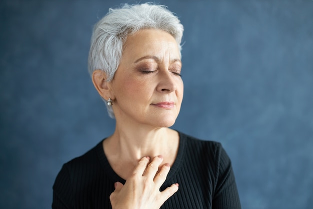 Primer plano de elegante atractiva mujer de mediana edad con canas y arrugas, cerrando los ojos y sonriendo, tocando el cuello, aplicando crema anti envejecimiento