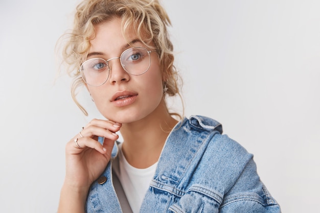 Primer plano elegante guapo de moda rubia blogger de ojos azules usar gafas transparentes chaqueta de mezclilla tocando la barbilla mirada pensativa cámara intrigada, interesada, de pie pared blanca