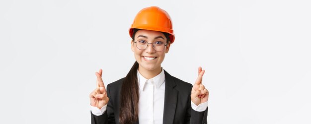 Primer plano de la emocionada y esperanzada ingeniera asiática gerente de construcción en casco de seguridad y traje de negocios suplicando saborear los dedos cruzados buena suerte y morder el labio tentador fondo blanco