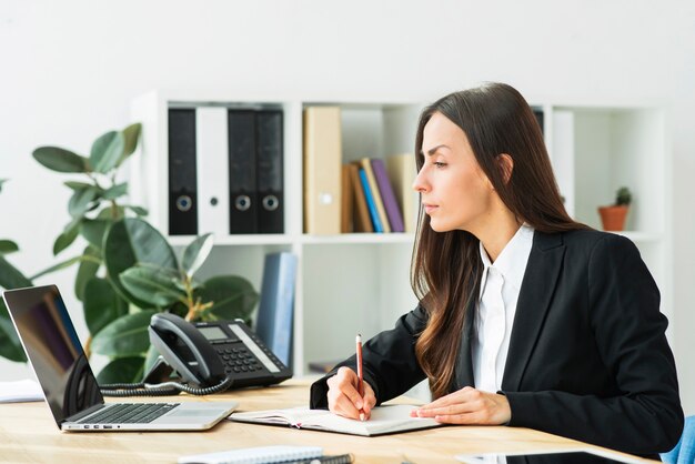 Primer plano de una empresaria mirando portátil escribiendo notas en diario con lápiz