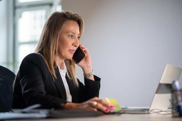 Primer plano de empresaria con teléfono. Mujer seria sentada en la mesa mirando la computadora portátil usando el teléfono hablando con socios sobre nuevos proyectos y apoyo financiero. Concepto de finanzas y crecimiento empresarial