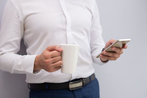 Primer plano del empresario de pie con taza de té con teléfono móvil