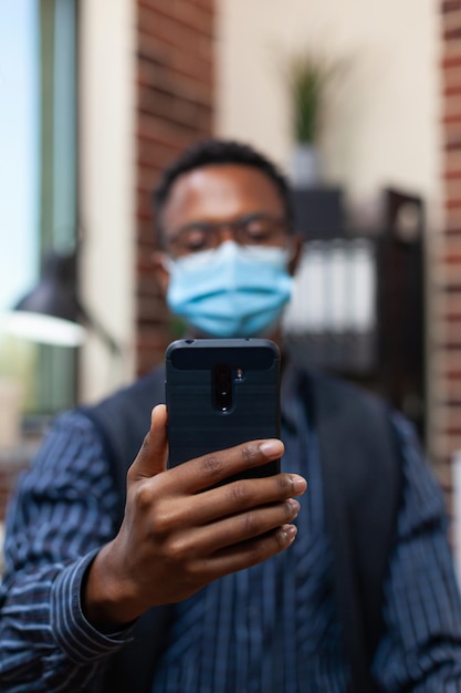 Primer plano de un empresario que sostiene un teléfono inteligente en una videoconferencia con una máscara protectora covid y auriculares inalámbricos. Vista detallada del teléfono inteligente de los empleados de inicio en una entrevista en línea.