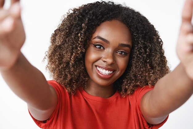 Primer plano de una encantadora mujer afroamericana tierna y femenina con cabello rizado en camiseta roja tirando de las manos y sosteniendo la cámara como abrazando o consolando a un amigo con una suave sonrisa en la cara