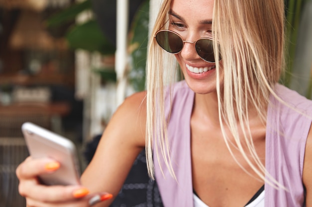 Primer plano de encantadora mujer alegre en gafas de sol con teléfono