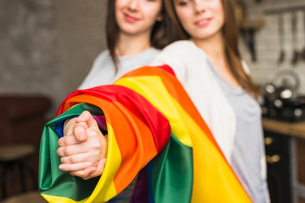 Primer plano de una encantadora pareja de jóvenes lesbianas tomándose las manos con la bandera de orgullo envuelto lgbt
