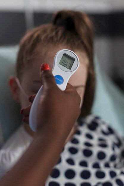 Primer plano de una enfermera afroamericana que mide la temperatura corporal de una niña enferma usando un termómetro. Personal médico del centro de salud infantil que examina la evolución de la enfermedad del niño enfermo bajo tratamiento