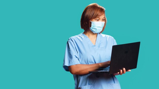 Primer plano de la enfermera conversando con el paciente en la computadora portátil durante la cita de control de telesalud en línea. Experto en atención médica feliz aislado sobre el fondo del estudio usando un aparato, usando mascarilla médica