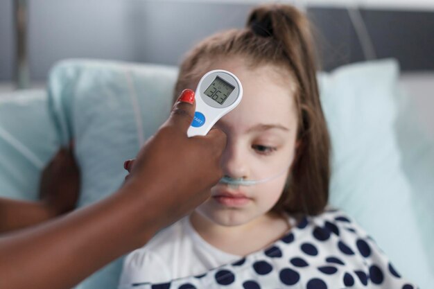 Primer plano de la enfermera médica que mide la temperatura corporal de la niña enferma usando un termómetro moderno. Enfermera midiendo la temperatura corporal del niño enfermo mientras se encuentra en la sala de recuperación de la clínica pediátrica.