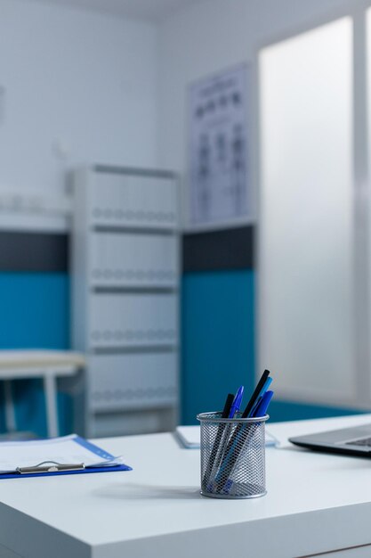 Primer plano del escritorio blanco de la oficina del médico del hospital con bolígrafos y portapapeles con una computadora portátil en la parte superior. Lugar de trabajo de la clínica moderna con gabinetes contemporáneos y diagrama médico en la pared