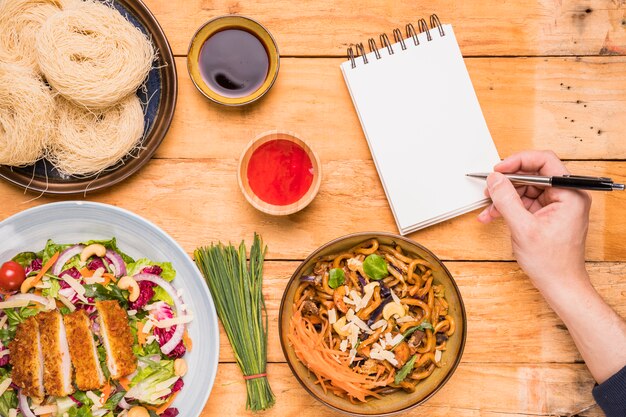 Primer plano de la escritura de una persona en la libreta con la pluma cerca de la comida tailandesa en la mesa de madera