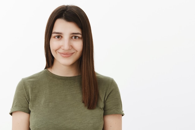 Foto gratuita primer plano de feliz encantadora y tierna linda joven morena de 20 años en camiseta verde oliva sonriendo tímida y con energía sintiéndose optimista mirando confiable sobre la pared blanca