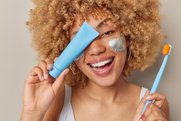 Primer plano de una feliz mujer europea de cabello rizado que aplica parches de belleza plateados sostiene un tubo de pasta de dientes sobre el cepillo de dientes de la cara que se ocupa de la belleza y la higiene oral aislada sobre el fondo gris