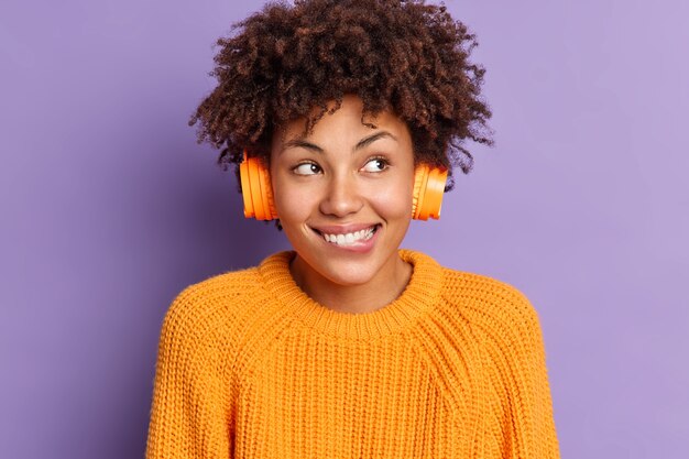 Primer plano de feliz mujer de piel oscura escucha música a través de auriculares, muerde los labios y disfruta de una agradable melodía vestida con poses de suéter naranja tejido