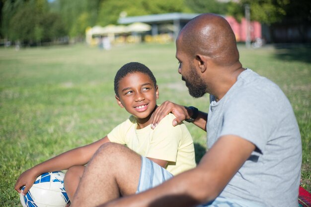 Primer plano de feliz papá afroamericano hablando con su hijo. Hombre guapo sentado en el suelo tocando el hombro del niño sonriente ambos mirando el uno al otro. Concepto de crianza, ocio y descanso activo juntos