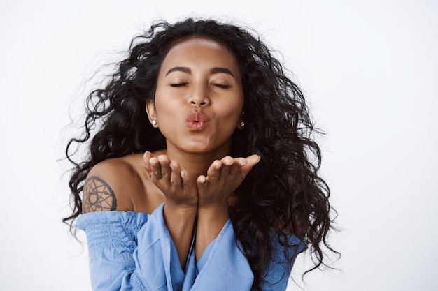 Primer plano femenino, tierna y romántica chica de pelo rizado con tatuajes en blusa azul, labios plegables, cerrar los ojos y enviar besos, soplar mwah coqueta, soporte pared blanca