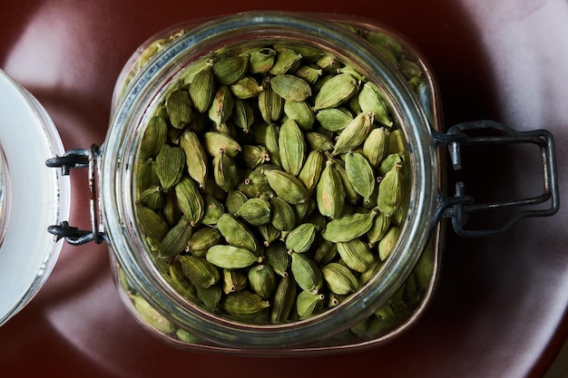 Primer plano de un frasco de vidrio lleno de vainas de cardamomo verde en un plato marrón. Vista superior.