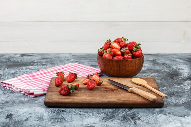 Primer plano de fresas y utensilios de cocina en tabla de cortar de madera con mantel de cuadros rojos y un tazón de fresas en mármol azul oscuro y fondo blanco de madera. horizontal