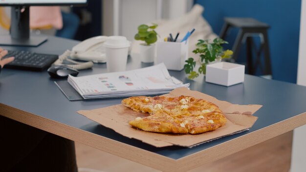 Primer plano del gerente ejecutivo tomando una rebanada de pizza comiendo frente a la computadora escribiendo gráficos financieros en la oficina de la empresa de inicio de negocios