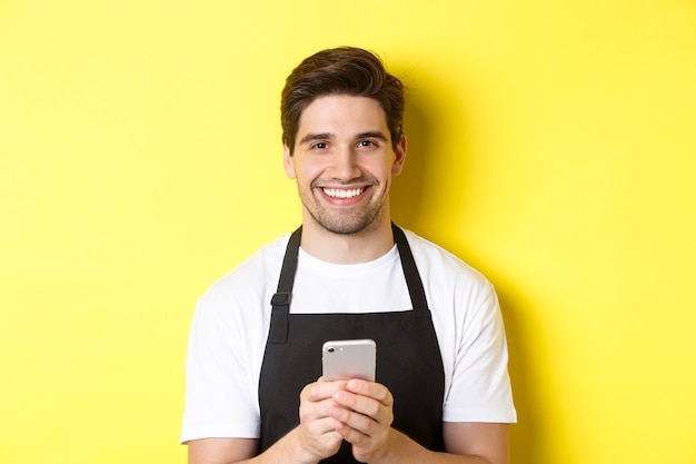 Primer plano de guapo barista enviando un mensaje en el teléfono móvil, sonriendo feliz, de pie sobre fondo amarillo