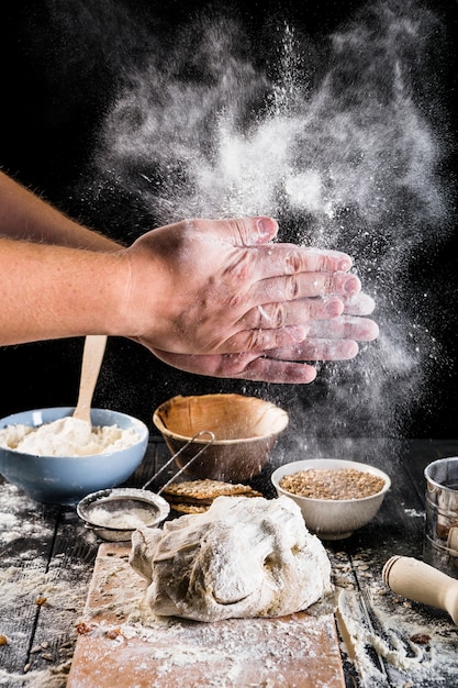 Primer plano de harina de polvo de la mano del panadero en la masa con los ingredientes en la mesa