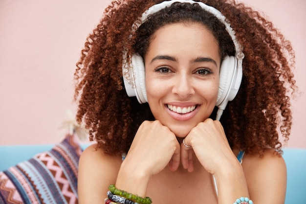 Primer plano de la hermosa joven africana encantada tiene una amplia sonrisa, disfruta de su melodía favorita en auriculares