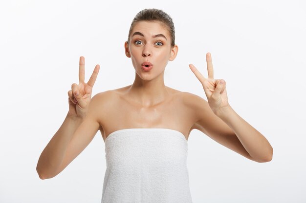 El primer plano de una hermosa joven con una toalla de baño que cubre sus senos en gris muestra el signo de dos dedos
