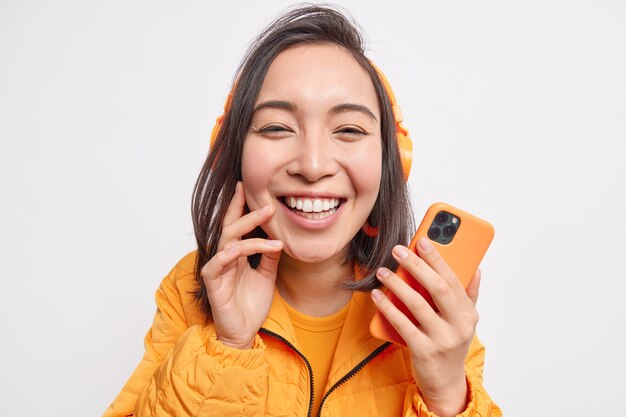 Primer plano de hermosa mujer asiática alegre sonríe ampliamente de deleite disfruta escuchando música favorita tiene teléfono móvil vestido con chaqueta naranja aislada sobre pared blanca.