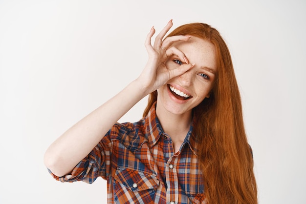 El primer plano de una hermosa mujer pelirroja sonriendo con dientes blancos mostrando un buen signo y luciendo feliz de pie sobre fondo blanco