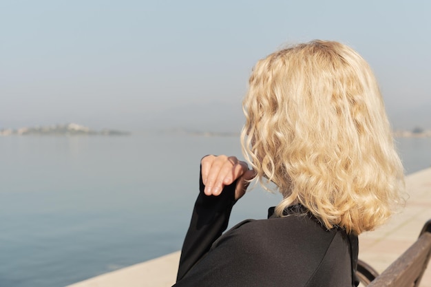 Primer plano de una hermosa mujer rubia adulta sentada en un banco y mirando el mar y la silueta azul del cielo de un espacio de mujer para la idea de texto para el fondo