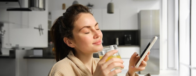 Foto gratuita el primer plano de una hermosa mujer sonriente bebe jugo de naranja fresco y disfruta de su mañana sostiene un teléfono móvil