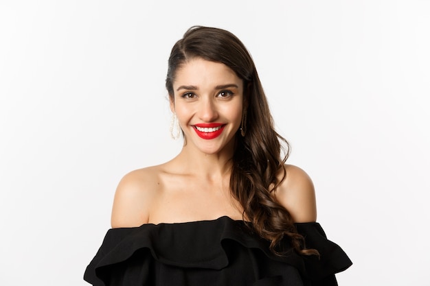 Primer plano de una hermosa mujer vestida para la fiesta en vestido negro, con maquillaje y lápiz labial rojo, sonriendo feliz a la cámara, fondo blanco.