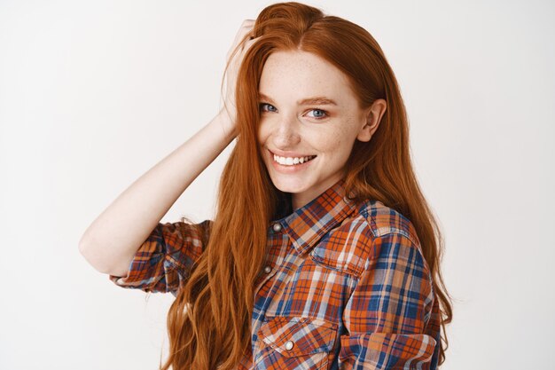 Primer plano de la hermosa pelirroja adolescente mujer tocando el cabello rojo natural, sonriendo con dientes blancos y mirando feliz, pared de estudio