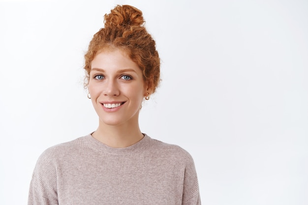 Primer plano, hermosa, tierna mujer pelirroja femenina con ojos azules, cabello rizado en moño, usar blusa de moda, mirar la cámara encantada y feliz, sonriendo complacida, de pie satisfecha sobre la pared blanca