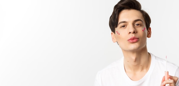 Primer plano de un hermoso hombre gay con brillo en los labios fruncidos de la cara después de aplicar el brillo de labios de pie