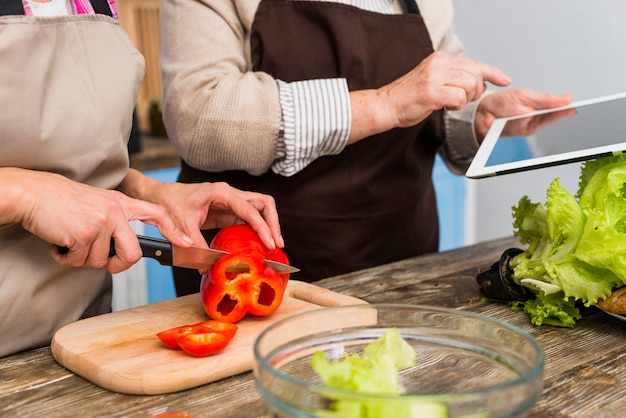 Primer plano de la hija de pie con su madre con tableta digital cortando pimiento con cuchillo