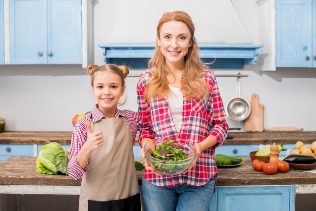 Primer plano de una hija sonriente que muestra el pulgar hacia arriba signo de pie con su madre sosteniendo un tazón de ensalada