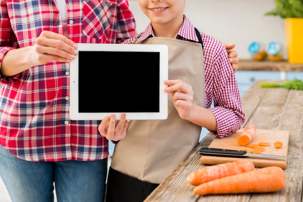 Primer plano de la hija y su madre mostrando tableta digital con zanahoria de mesa