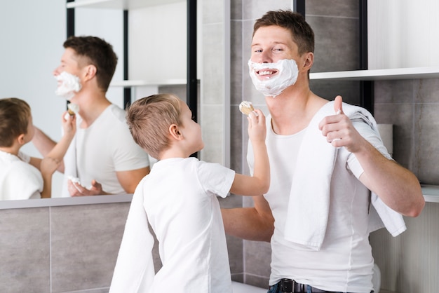 Primer plano de un hijo ayudando a su padre para afeitarse la barba mostrando el pulgar hacia arriba signo