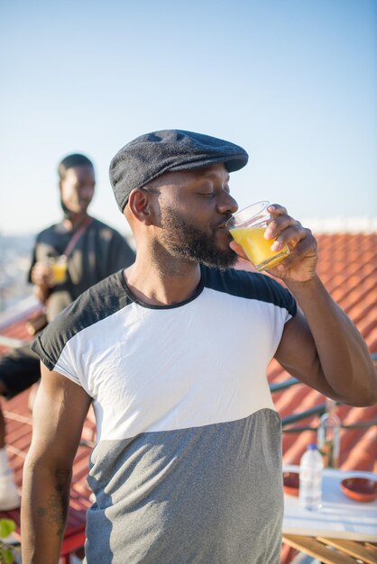 Primer plano de un hombre afroamericano bebiendo jugo en la azotea. Hombre guapo con gorra negra de pie bebiendo jugo y su amigo sentado en la valla detrás de él. Pasar el tiempo libre, el ocio, el concepto de relaciones.