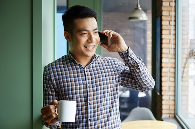 Primer plano de un hombre asiático con una taza de café mirando por la ventana mientras habla por teléfono