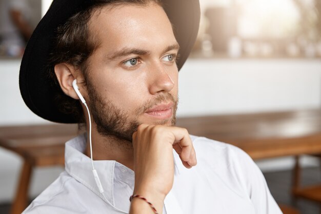 Primer plano de un hombre atractivo y elegante con barba que parece pensativo, escuchando música en línea en auriculares blancos usando algún dispositivo electrónico mientras espera a su novia en la cafetería