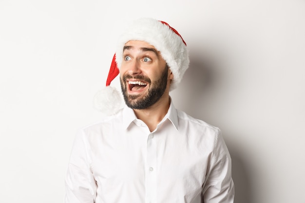 Foto gratuita primer plano de hombre barbudo asombrado con gorro de papá noel, mirando a la izquierda con entusiasmo, oferta promocional de navidad, de pie