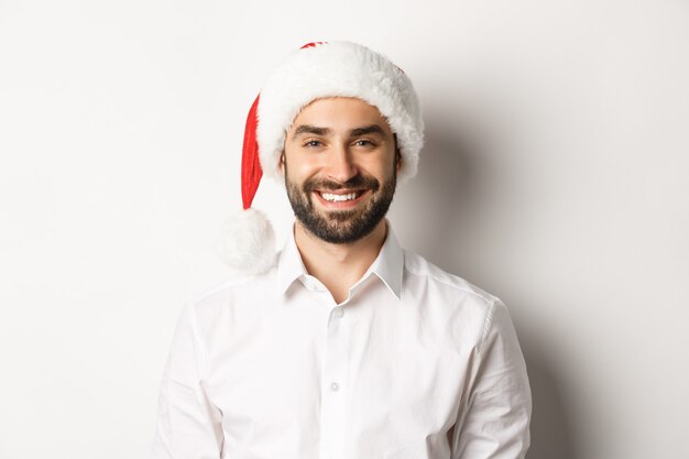 Primer plano de hombre barbudo feliz celebrando la Navidad, vistiendo gorro de fiesta de santa y sonriendo, de pie