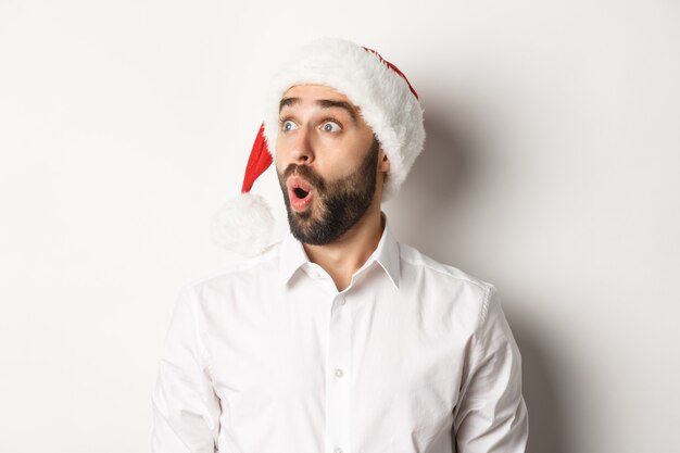 Primer plano de hombre barbudo sorprendido con gorro de Papá Noel, mirando a la izquierda con cara de sorpresa, oferta promocional de Navidad, de pie