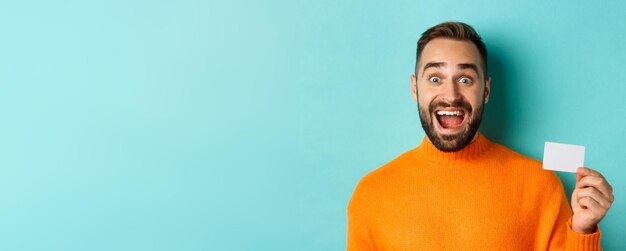 Primer plano de hombre caucásico emocionado mostrando su tarjeta de crédito sonriendo y mirando asombrado de pie en oran